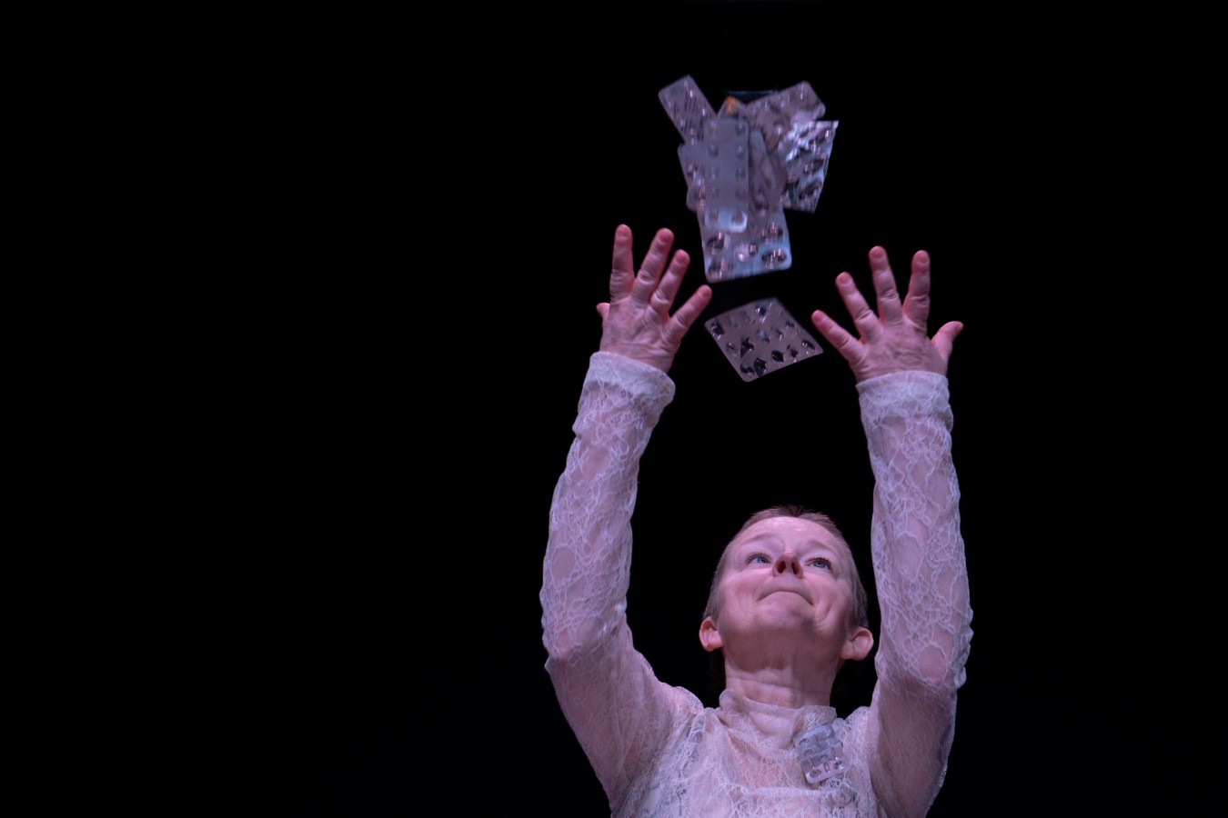 Katarzyna Żeglicka, Dzikorosła; fot. Grzegorz Krzysztofik. Katarzyna Żeglicka ubrana w białą koronkę, podrzuca w górę srebrne blistry po pigułkach. Widzimy tylko jej podniesione w górę ramiona i głowę. Ma zaciśnięte usta, patrzy na opakowania. Jasna postać kontrastuje z czarnym tłem.