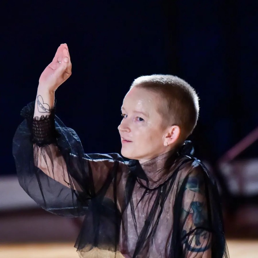 Katarzyna Żeglicka, PokaZ; fot. Erdelji Marija. Katarzyna Żeglicka jest ubrana w luźny kostium z czarnego tiulu, spod którego prześwituje jej nagie ciało. W centrum zdjęcia są jej klatka piersiowa, ogolona głowa i podniesiona w górę prawa ręka. Żeglicka jest ustawiona profilem, ale patrzy w stronę obiektywu, lekko się uśmiecha. Podniesioną w górę rękę zdobią tatuaż w kształcie serca z napisem w środku oraz srebrna obrączka. 