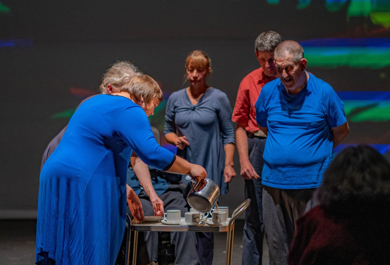 Dress rehearsal for Mam (2023): ‘Pouring the Tea’. From left to right the performers are Arlene James, Karen Rush, Agnieszka Michalik, Sam Evans and Adrian Jones. Five people standing around a small table: three women and two men. They are wearing everyday clothes in different colours. One of the women (in a blue dress) is pouring some beverage from a jug into the cups that are standing on the table.