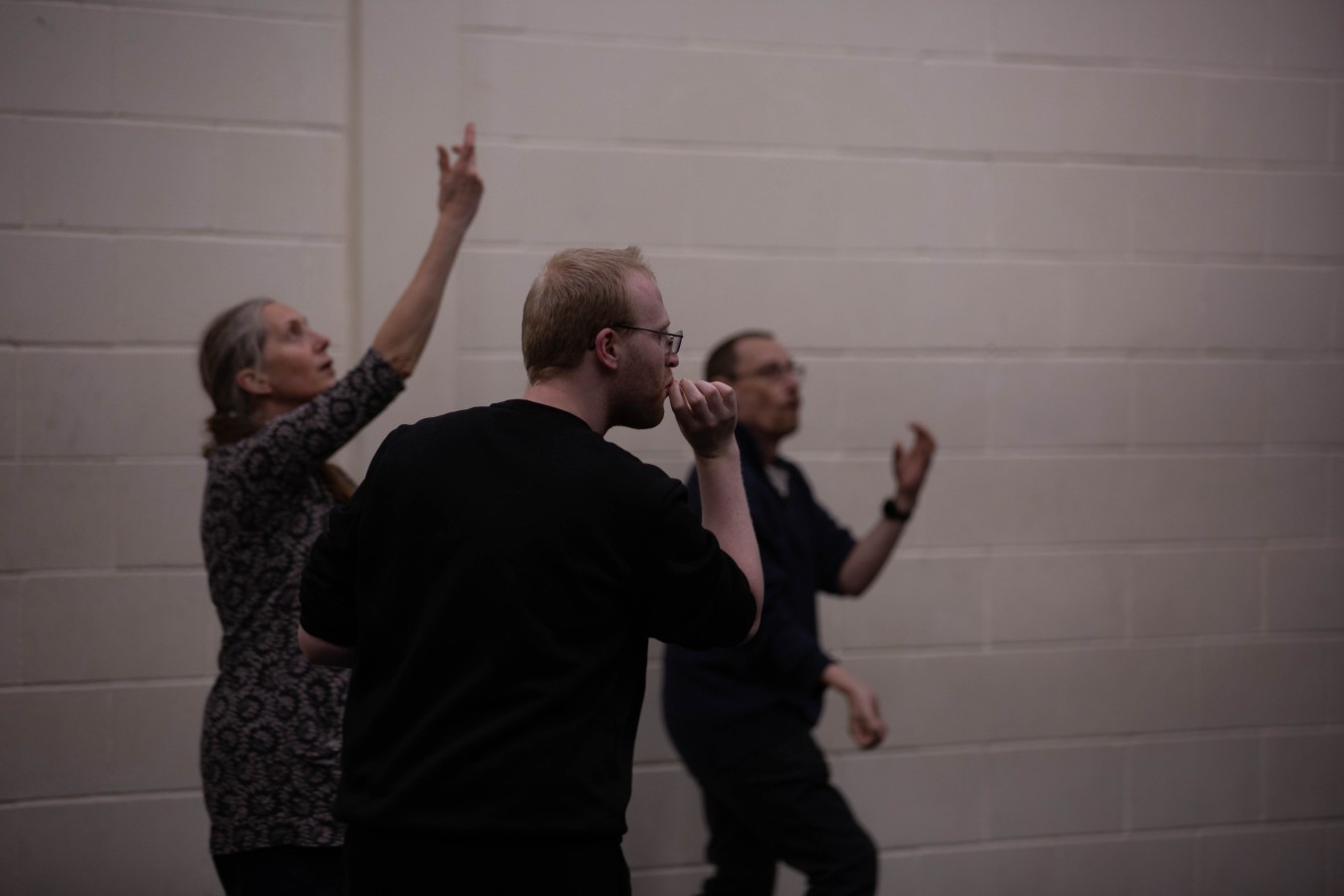 In My Younger Years – rehearsal in the section 'Juggling': Sam Cleeton, Karen Rush, David Bartholemew-Biggs. Three persons, one woman and two men, in dark clothes against a white brick wall. Two of them pretend to be juggling, the other man puts his fingers to his mouth as if he were smoking.