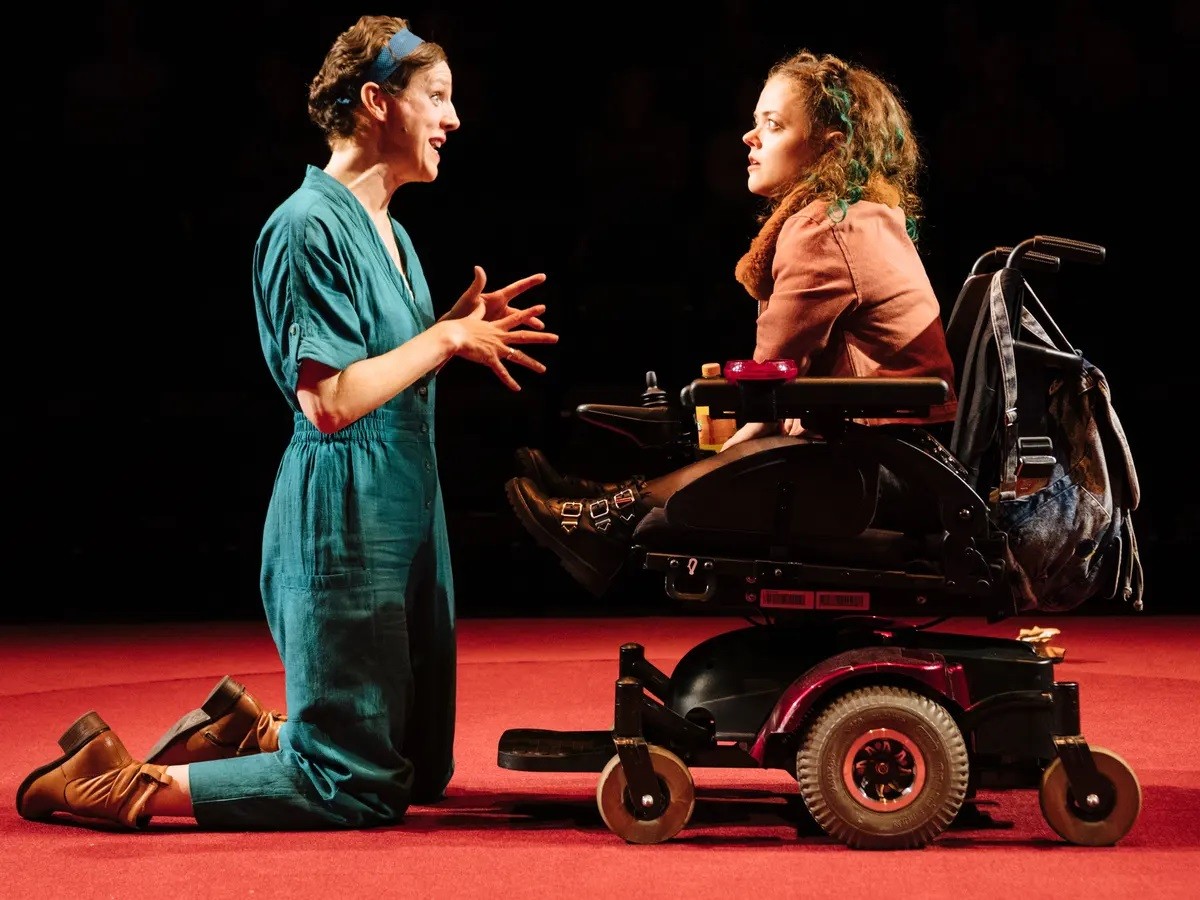 Dorfman Theatre w Londynie, All of Us: Francesca Martinez (Jess), Francesca Mills (Poppy). Two women facing each other, sideways to the viewer. The tall woman on the left is kneeling. She is dressed in a green jumpsuit and brown shoes. She seems to be saying something funny to the next woman. The woman on the right is listening with attention. She is dressed in a brown jumper and black shoes. She is sitting in an electric wheelchair. The floor is red, and the background is black.
