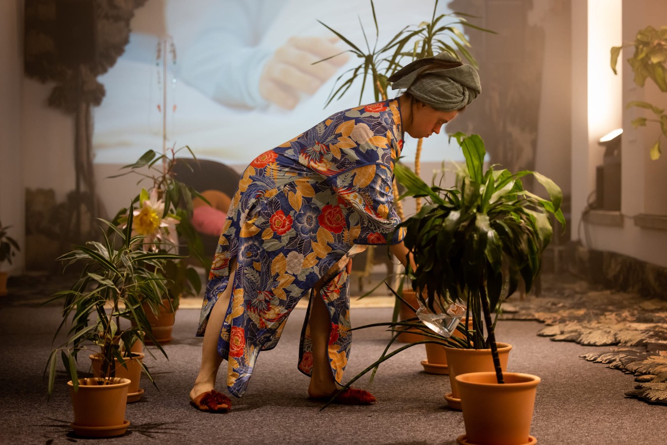 Teatr 21, Nie jestem rośliną. Strumień świadomości: Aleksandra Skotarek; fot. Alicja Szulc   Teatr 21, Nie jestem rośliną. Strumień świadomości: Aleksandra Skotarek; fot. Alicja Szulc. Aleksandra Skotarek podlewa kwiaty doniczkowe, które stoją w dwóch rzędach, po jednym z każdej strony. Usytuowana na środku kadru aktorka ma na sobie szlafrok we wzory kwiatów i liści oraz zrobiony z ręcznika turban.