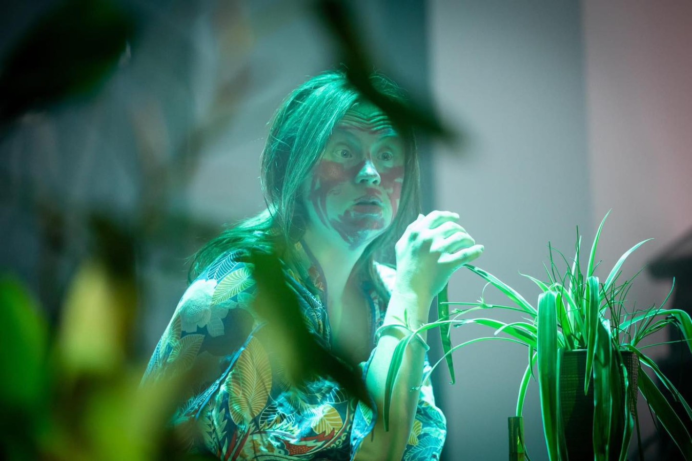 Teatr 21, Nie jestem rośliną. Strumień świadomości: Aleksandra Skotarek; fot. Alicja Szulc Teatr 21, Nie jestem rośliną. Strumień świadomości: Aleksandra Skotarek; fot. Alicja Szulc. Aleksandra Skotarek siedzi wśród roślin o długich, wąskich, zielonych liściach. Aktorka ma na sobie szlafrok we wzory kwiatów i liści, jej twarz jest nieregularnie pomalowana na czerwono. Patrzy w bok, ma szeroko otwarte oczy i zmarszczone czoło, prawa dłoń jest uniesiona na wysokość twarzy. Wygląda na zdziwioną lub zaskoczoną.