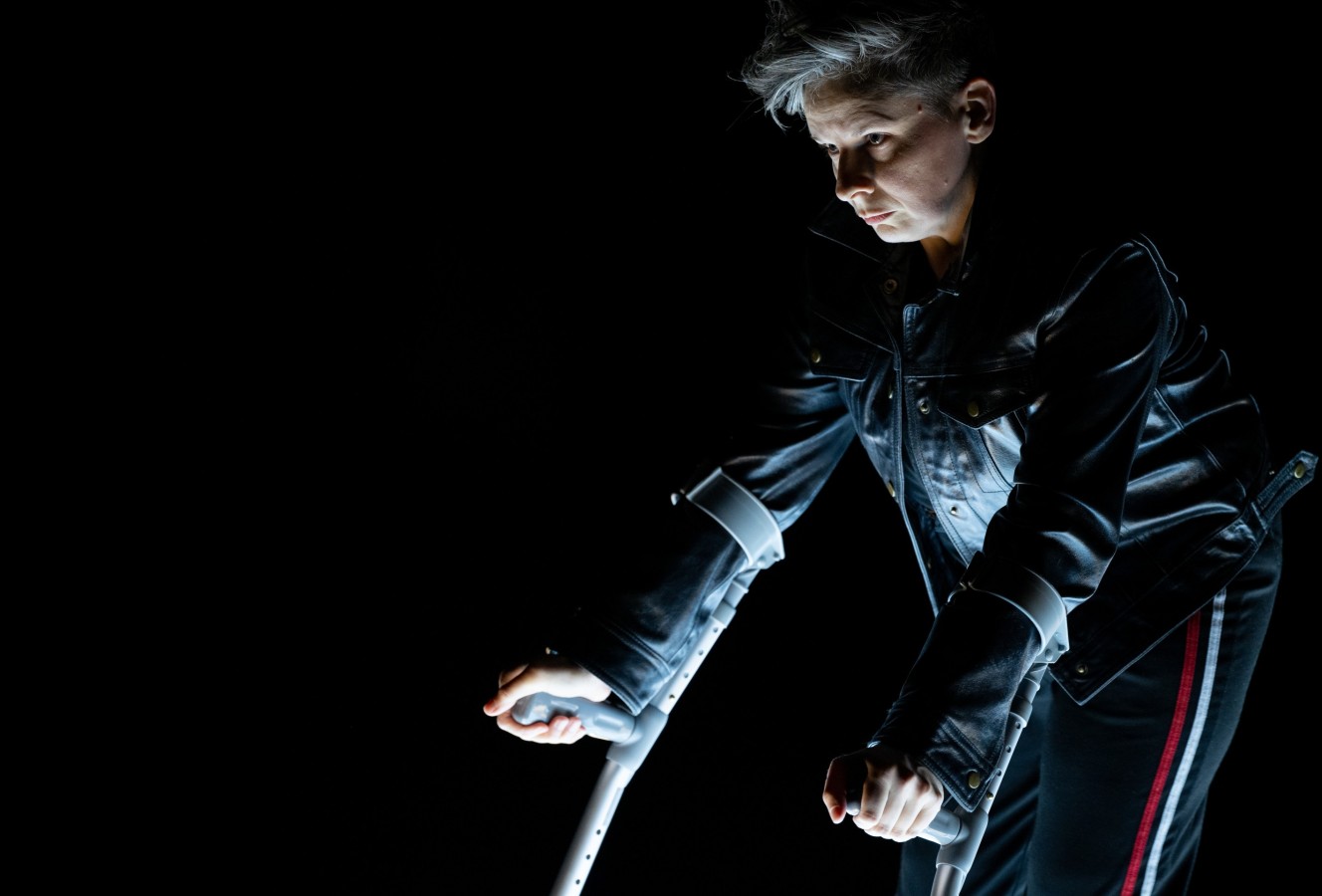 Thank You Very Much: Claire Cunningham; photo: ©Lars Opstad. A close-up of Claire Cunningham, a middle-aged person with short blond hair and shaved sides. Lit from below, she stands in profile, against a black background, leaning forward on her grey elbow crutches. Cunningham is dressed in a black leather jacket and black sweatpants with red and white side stripes. Her face is composed as she keeps looking at something intently. 