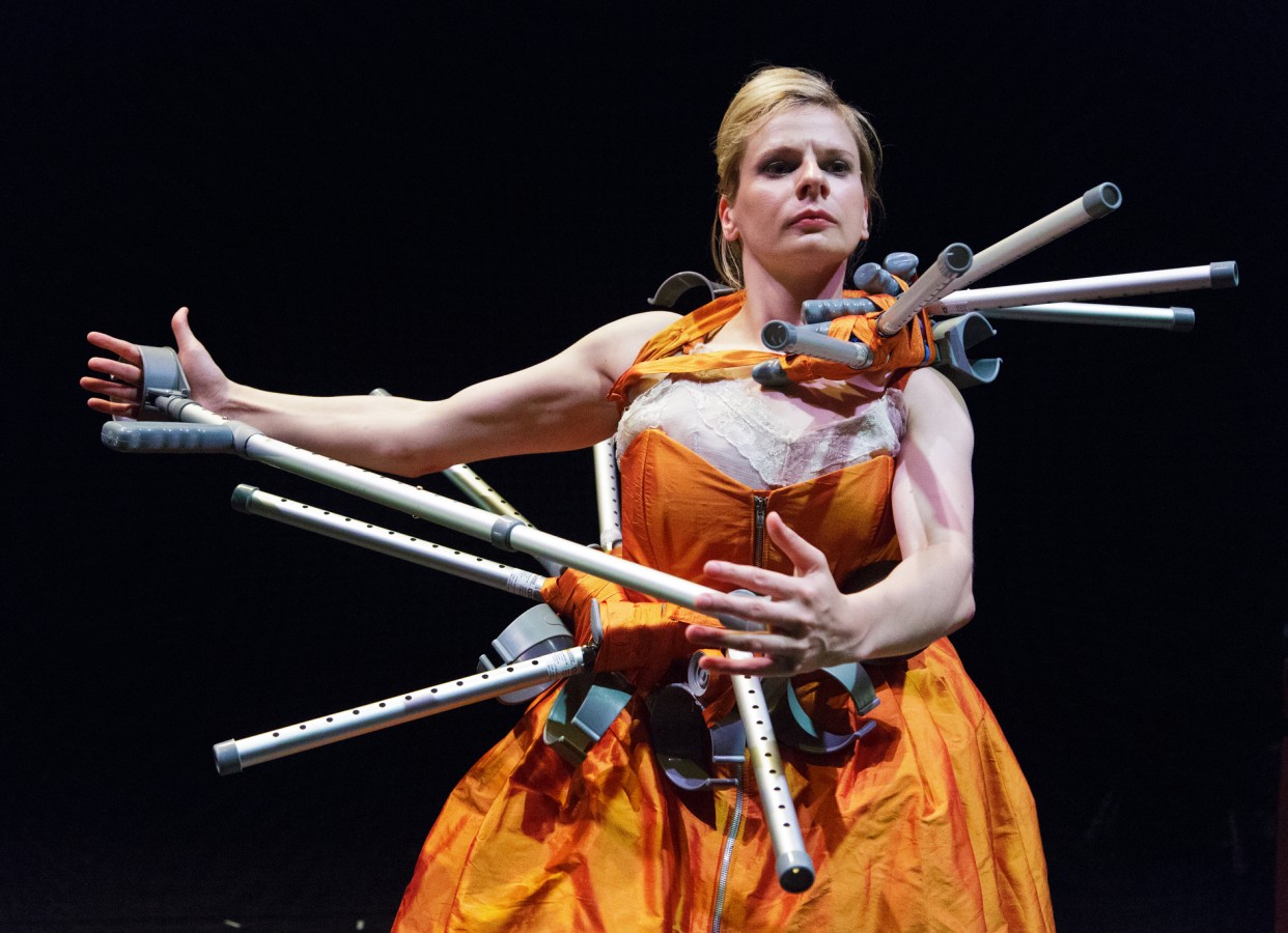 Ménage à Trois: Claire Cunningham; photo: ©Kenny Mathieson. Claire Cunningham is shown en face. She stands in front of the camera, holding her head high. Cunningham has smoothly styled hair and is dressed in an orange silk dress, which resembles female gowns of the past. At the waist and close to the neck, the dress is decorated with short spike-like crutches. At waist level, Cunningham holds one of her regular crutches horizontally in her muscular arms: the cuff in one hand and the ferrule in the other. 