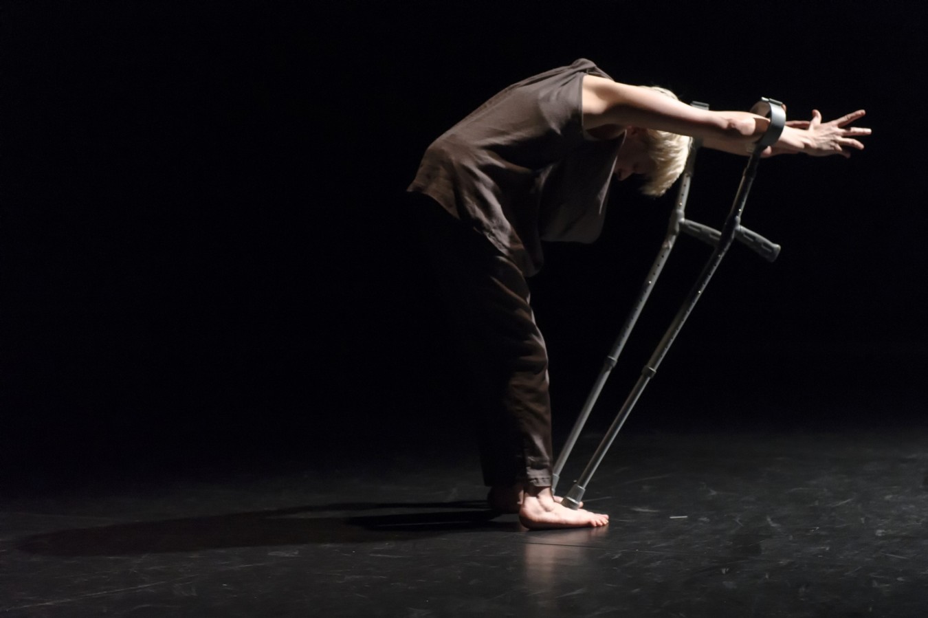 Give Me a Reason to Live: Claire Cunnigham; photo: ©Ben Nienhuis. Cunningham stands on a bare black stage. We see her from the side. She is dressed in long black trousers and a black vest. The ferrules of her crutches rest on her bare feet. Cunningham has thrust her hands though the crutch cuffs and lowered her head. The crutches tilt forward.