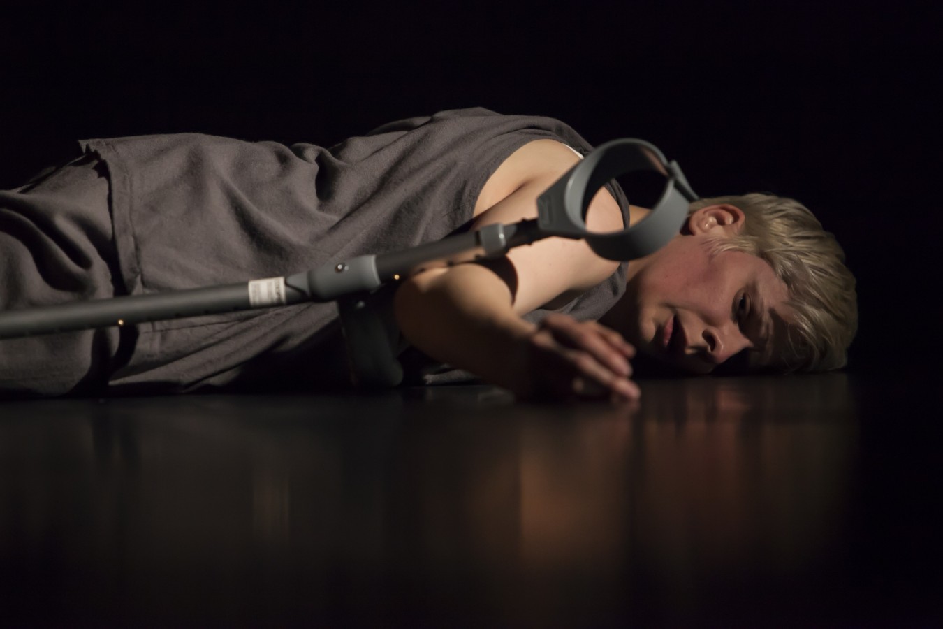 Give Me a Reason to Live: Claire Cunnigham; photo: ©Maria Falconer. A close-up of Cunningham’s upper body, which lies on a black stage and faces the viewer. She is dressed in long black trousers and a black vest. Her facial expression – half-closed eyes and slightly parted lips – suggests exhaustion. Her right arm is extended towards the viewer. One of her grey crutches rests on that arm. 