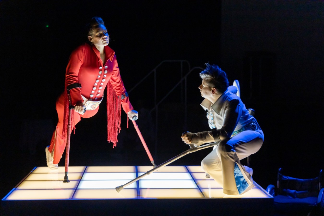 Thank You Very Much: Vicky Malin, Claire Cunningham; photo: M. Kaczyński ©CK ZAMEK. Dressed in a white Elvis costume, Cunningham stands on a podium with a grid of white lights. Supported by her crutches, she crouches down and rests her left arm on the knee. She is looking at the person in front of her, Tanja Erhart – a one-legged middle-aged woman dressed in a red Elvis outfit. Returning Cunningham’s gaze, Erhart leans forward on her red glitter crutches. This looks like either collaboration or a stage duel