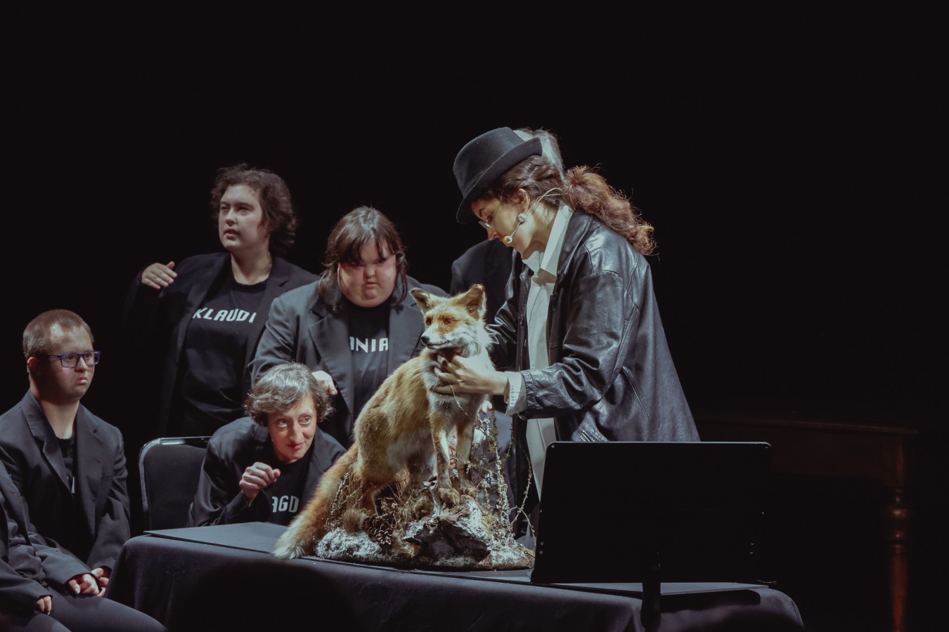Ośrodek Dokumentacji Sztuki Tadeusza Kantora Cricoteka, Festiwal Kultury Wrażliwej, Lekcja anatomii – rekonfiguracja; fot. Magda Rymarz. Na czarnym stole stoi wypchany lis, wokół którego zgromadziła się grupa sześciu ubranych na czarno osób. Jedna z nich, w białej koszuli i z kapeluszem na głowie, dotyka lisa, trzy przyglądają się z zaciekawieniem jej działaniom, jedna patrzy w bok, w stronę niewidocznej w kadrze publiczności, a szósta osoba jest prawie niewidoczna