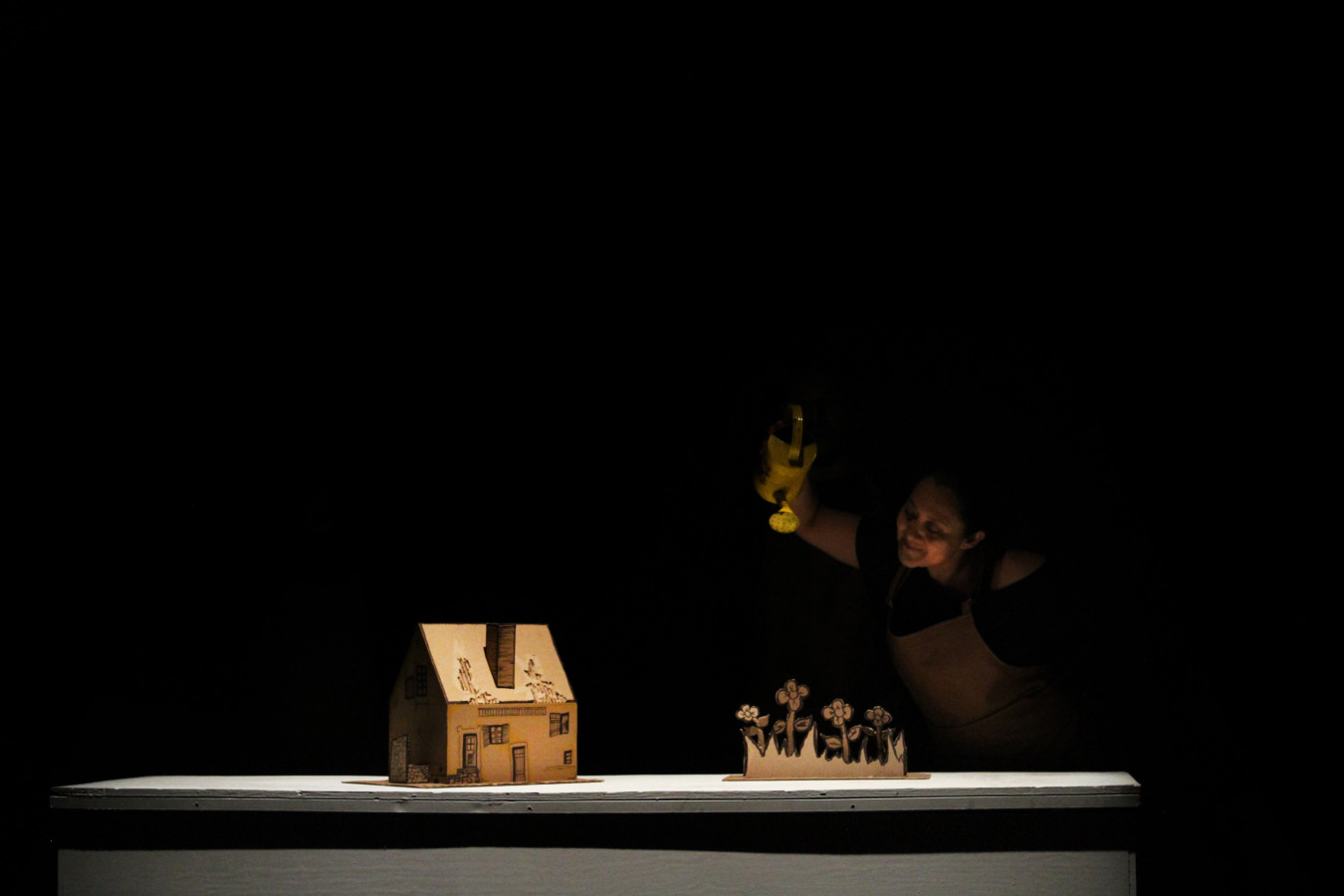 Teatro Ciego MX, La mirada del inventor ciego: Susana Romero; fot. Héctor Ortega. On a dark stage, there is a bright table with two objects: a small cardboard model of a house on the left and a flower cutout on the right. Behind the table, a woman wearing a brown apron holds a yellow watering can. She leans forward and makes a gesture as if watering the flowers. The background is completely black, highlighting the contrast with the lit table.