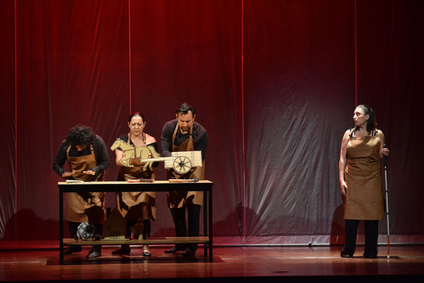 Teatro Ciego MX, La mirada del inventor ciego: Marco Antonio Martínez, Susana Romero, Juan Carlos Sáenz, Erika Bernal; fot. Héctor Ortega. In front of a red curtain, four actors in brown aprons stand on stage. Three are on the left at a table, one holding a horse cut-out, while the others gesture as if working. On the right, a woman with a white cane faces the audience but looks to the side, watching them. The stage is lit with warm red light.