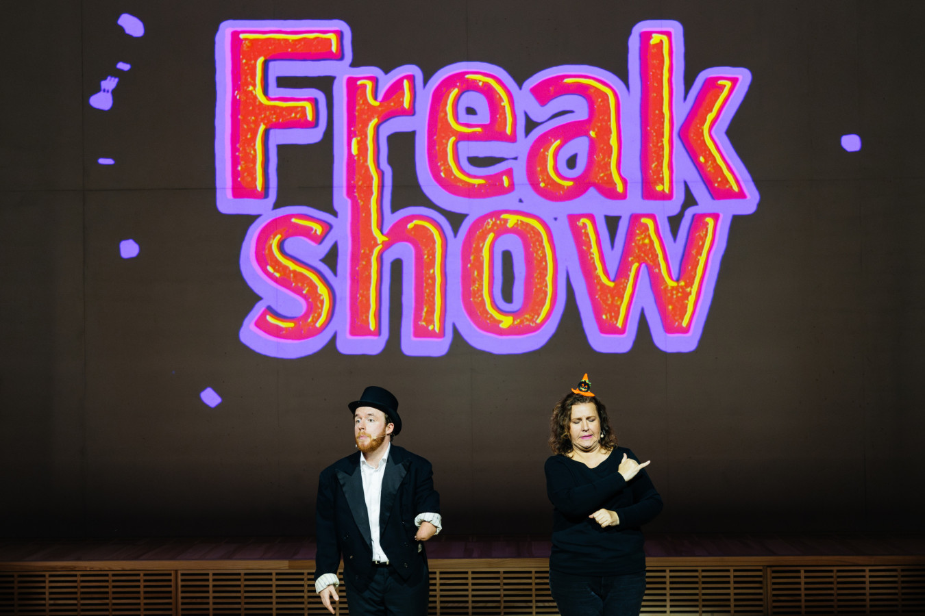 Fundacja Inqubator Teatralny, Freak Show: Filip Pawlak, Katarzyna Głozak; fot. Dominika Jaruga Fundacja Inqubator Teatralny, Freak Show: Filip Pawlak, Katarzyna Głozak; fot. Dominika Jaruga. Na zdjęciu widać dwie osoby stojące na scenie na tle dużego, kolorowego napisu „Freak show”. Po lewej: Filip Pawlak w czarnym fraku i cylindrze, z rudą brodą; jego, odsłonięta dzięki podwiniętym rękawom, prawa ręka jest znacznie krótsza od lewej. Patrzy wymownie i pytająco w stronę publiczności. Po prawej: Katarzyna Głozak, ubrana na czarno, ma małą czapeczkę halloweenową, wykonuje znak języka migowego.